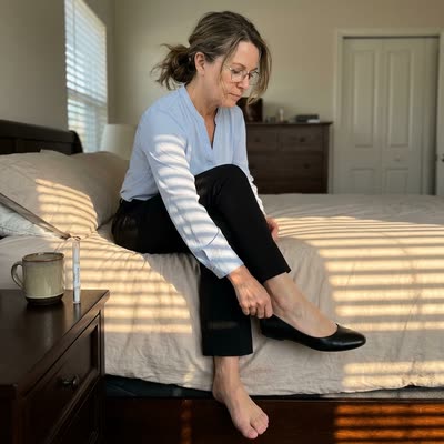Morning bedroom — Karen slipping on a work flat with the NailScience pen on the nightstand behind her