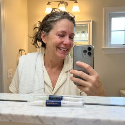 Bathroom mirror selfie — Linda smiling with tears of relief, NailScience Step 2 pen on marble vanity